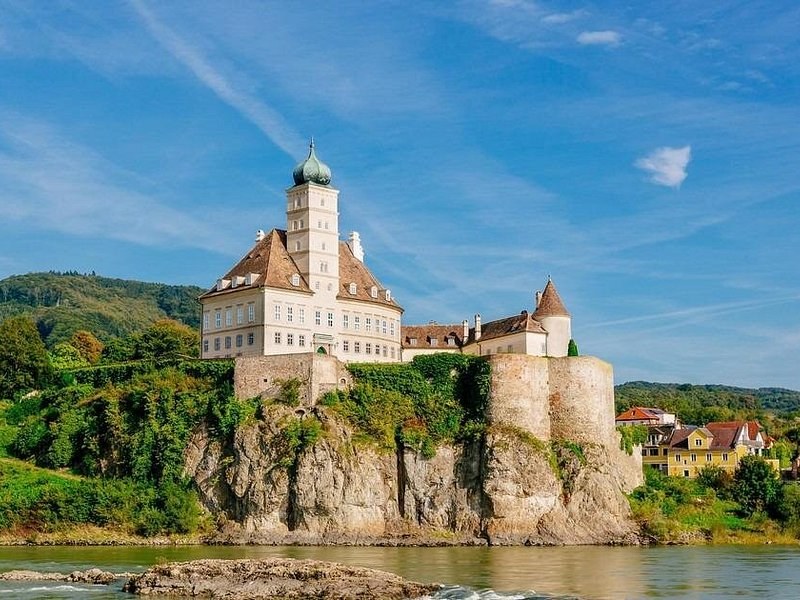 A white castle with an onion-domed tower on a rock above the Danube